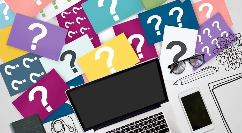 laptop em uma mesa cercada por folhas coloridas com pontos de interrogação, simbolizando a seleção de palavras-chave